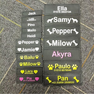 May include: A selection of black rectangular name patches with white, yellow, and pink text. The patches feature names like "Jack", "Ella", "Samy", and "Paulo", alongside phone numbers and symbols including paw prints, hearts, and bones. These are likely pet name tags.