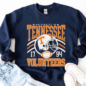 May include: A navy blue crewneck sweatshirt featuring a graphic print that reads University of Tennessee Volunteers with the numbers 17 and 94. The design includes a white football helmet with an orange T logo and a football in orange and white.