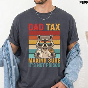 Puede incluir: Camiseta gris oscuro con un diseño retro. Presenta un mapache con gafas de sol, sosteniendo una bebida y papas fritas. El texto dice "DAD TAX MAKING SURE IT'S NOT POISON" en rojo, naranja, amarillo y verde.