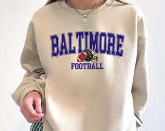 May include: A sand-coloured sweatshirt featuring the word "BALTIMORE" in blue block letters, with "FOOTBALL" beneath. A football and helmet graphic is centered below "BALTIMORE". The sweatshirt has long sleeves and a crew neck.