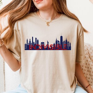 May include: A sand-colored t-shirt featuring a blue and red graphic of the New York City skyline with the word "INDIVISIBLE" in red. The shirt is made of a soft material and has a classic crew neck.