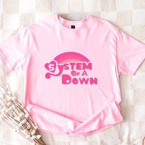May include: Pale pink t-shirt featuring the text "System Of A Down" in a pink and magenta rainbow design. The short-sleeved shirt is displayed on a white and beige checkered surface. The shirt is a light pink colour.