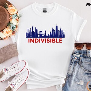 May include: White t-shirt featuring a blue skyline graphic with the Statue of Liberty and the Brooklyn Bridge. The word "INDIVISIBLE" is printed in red below the skyline. The shirt is styled with denim shorts, sunglasses, and white sneakers.