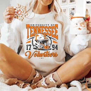 May include: White sweatshirt with orange and black graphic of a football helmet and the words "University of Tennessee 17 94 Volunteers". The person is holding a snowflake cookie and a Santa mug.