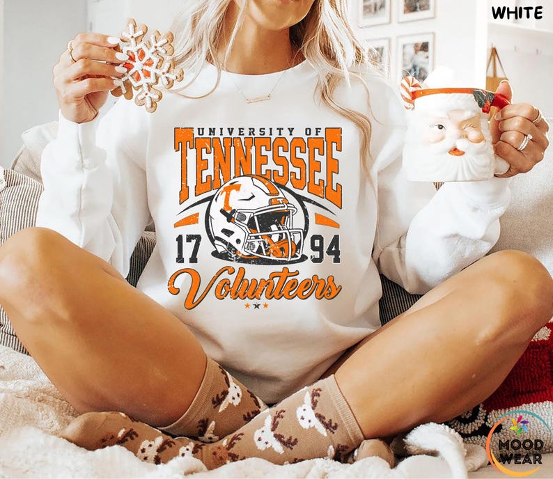 May include: White sweatshirt with orange and black graphic of a football helmet and the words "University of Tennessee 17 94 Volunteers". The person is holding a snowflake cookie and a Santa mug.
