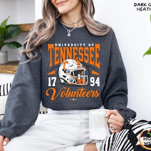May include: Dark gray heather sweatshirt featuring the University of Tennessee Volunteers logo in orange and white. The design includes a football helmet, the years 17 and 94, and the word "Volunteers".