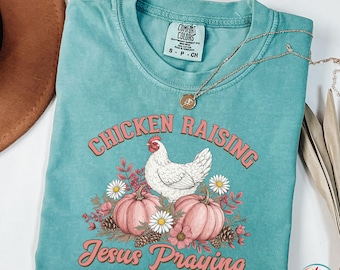 May include: A turquoise Comfort Colors t-shirt with the words "Chicken Raising Jesus Praying" in pink script. The graphic features a white chicken, pink pumpkins, flowers, and the brand's label. A gold necklace is draped across the shirt.