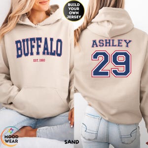 May include: Sand-colored hoodie with the word "BUFFALO" in navy blue block letters with a red outline. The hoodie also features the text "EST. 1960". The back of the hoodie displays the name "ASHLEY" and the number "29" in navy blue with a red outline.