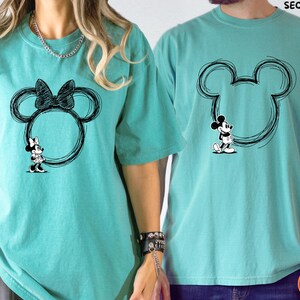 May include: Two turquoise t-shirts with black Mickey Mouse and Minnie Mouse designs. The Minnie Mouse shirt features a bow and a Minnie Mouse figure. The Mickey Mouse shirt has a Mickey Mouse figure. Both designs are inside a circular outline.