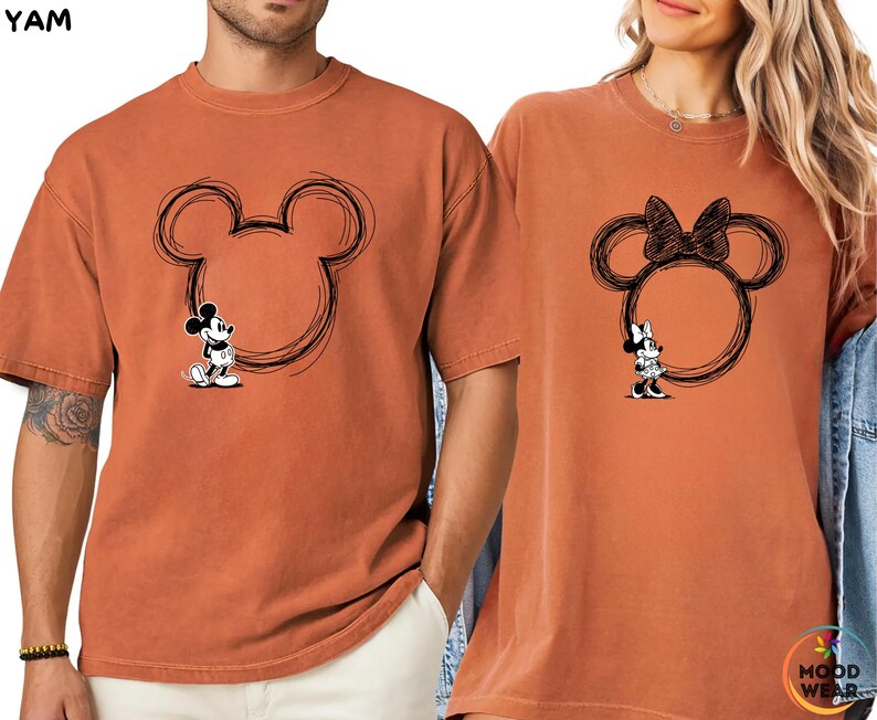 May include: Two rust-colored t-shirts featuring Mickey and Minnie Mouse designs. One shirt has a Mickey Mouse graphic inside a sketched outline of Mickey's head. The other shirt has a Minnie Mouse graphic inside a sketched outline of Minnie's head with a bow.