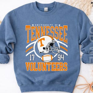 May include: A blue crewneck sweatshirt featuring a graphic print that reads University of Tennessee Volunteers with a football helmet and a football. The text includes the numbers 17 and 94. The design is in orange and white on the front of the garment.