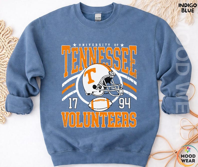 May include: A blue crewneck sweatshirt featuring a graphic print that reads University of Tennessee Volunteers with a football helmet and a football. The text includes the numbers 17 and 94. The design is in orange and white on the front of the garment.