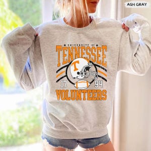 May include: A light gray crewneck sweatshirt featuring a distressed orange and black graphic that reads University of Tennessee Volunteers with the numbers 17 and 94, a football helmet with a T logo, and a football centered on the front chest.