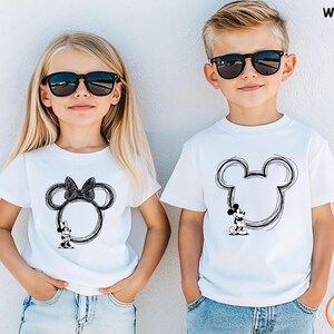 May include: Two children wearing white t-shirts with black Mickey Mouse and Minnie Mouse designs. The Mickey Mouse design is on the right, and the Minnie Mouse design is on the left. Both children are wearing black sunglasses and blue jeans.