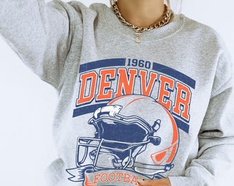 May include: An athletic grey crewneck sweatshirt featuring a graphic print with the text 1960 DENVER FOOTBALL. The design includes a stylised football helmet in orange and navy blue, with the text displayed in bold orange and navy blue lettering.