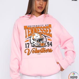 May include: Light pink hoodie with a graphic featuring "University of Tennessee" in orange block letters, a football helmet, and the text "Volunteers 17 94". The hoodie has a drawstring hood and a front pocket.
