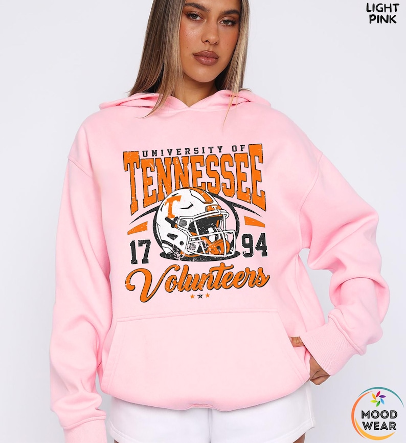 May include: Light pink hoodie with a graphic featuring "University of Tennessee" in orange block letters, a football helmet, and the text "Volunteers 17 94". The hoodie has a drawstring hood and a front pocket.