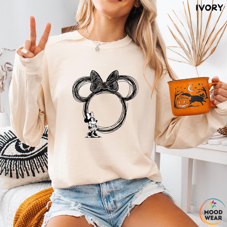 May include: Ivory-colored sweatshirt featuring a black sketch of Minnie Mouse's head and bow. The design includes a Minnie Mouse figure. The word "IVORY" is printed at the top. A Halloween-themed orange mug is held in the hand.