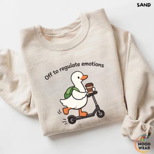 Peut inclure: Sweat-shirt couleur sable avec un dessin d'un canard blanc sur un scooter noir, tenant une tasse de café et un sac à dos vert. Le texte "Off to regulate emotions" est au-dessus du canard. Le sweat-shirt a un col rond et des manches longues.