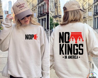 May include: Sand-colored sweatshirt with the text "NOPE" and a red crown graphic on the front. The back of the sweatshirt features the text "NO KINGS IN AMERICA" with a red crown graphic. The model is wearing a baseball cap.