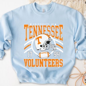 May include: A light blue crewneck sweatshirt featuring a graphic print with the text University of Tennessee Volunteers, the numbers 17 and 94, a football helmet with a T logo, and a football in orange and white with a distressed texture.