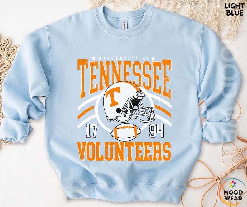 May include: A light blue crewneck sweatshirt featuring a graphic print with the text University of Tennessee Volunteers, the numbers 17 and 94, a football helmet with a T logo, and a football in orange and white with a distressed texture.