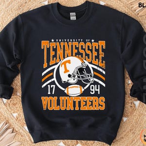 May include: A black long-sleeve sweatshirt featuring a graphic print with the text University of Tennessee Volunteers, the numbers 17 and 94, a white football helmet with an orange T logo, and a football in orange and white distressed lettering.