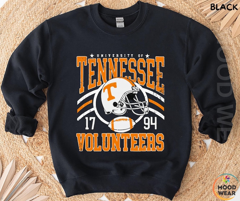 May include: A black long-sleeve sweatshirt featuring a graphic print with the text University of Tennessee Volunteers, the numbers 17 and 94, a white football helmet with an orange T logo, and a football in orange and white distressed lettering.