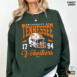 May include: Forest green sweatshirt with the text "University of Tennessee" in orange, along with a football helmet graphic. The numbers "17" and "94" are on either side of the helmet, and the word "Volunteers" is below.