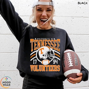 May include: A black long-sleeve sweatshirt featuring a graphic print that reads University of Tennessee Volunteers with the numbers 17 and 94, centered around an illustration of a football helmet and a football in orange, white, and black.