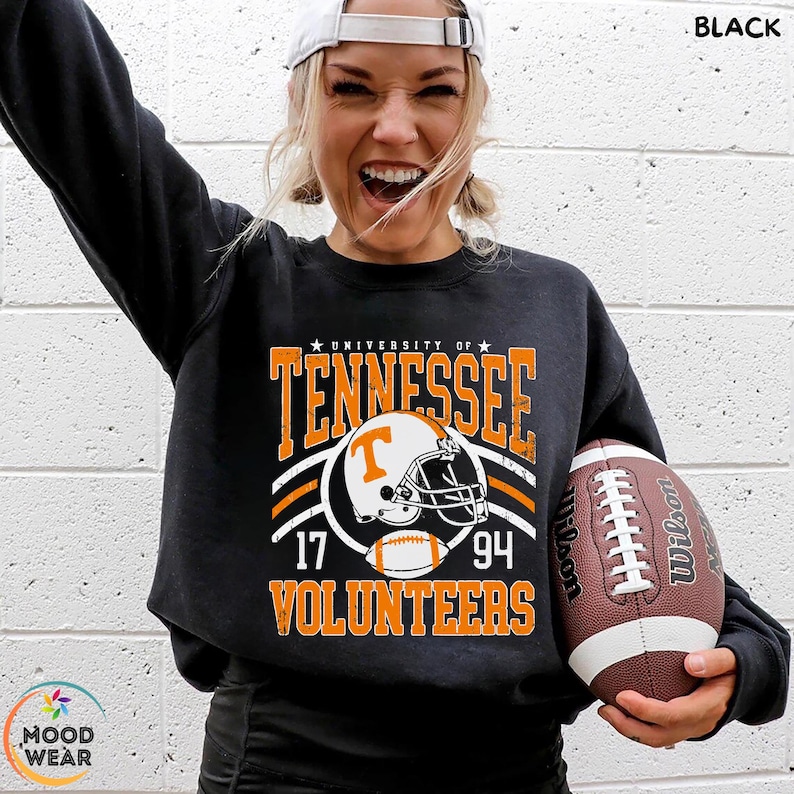 May include: A black long-sleeve sweatshirt featuring a graphic print that reads University of Tennessee Volunteers with the numbers 17 and 94, centered around an illustration of a football helmet and a football in orange, white, and black.