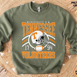 May include: A military green crewneck sweatshirt featuring a graphic print that reads University of Tennessee Volunteers with the numbers 17 and 94. The design includes a white and orange football helmet with a T logo and a football graphic.