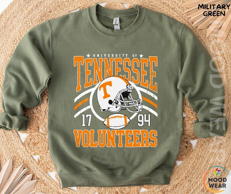 May include: A military green crewneck sweatshirt featuring a graphic print that reads University of Tennessee Volunteers with the numbers 17 and 94. The design includes a white and orange football helmet with a T logo and a football graphic.