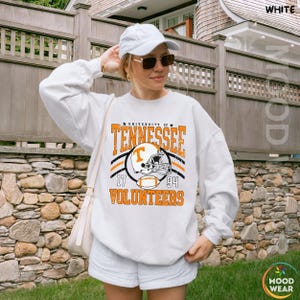 May include: A white long-sleeve sweatshirt featuring a graphic print that reads University of Tennessee Volunteers 1794 with an illustration of a football helmet and a football. The sweatshirt is paired with a white baseball cap and sunglasses.