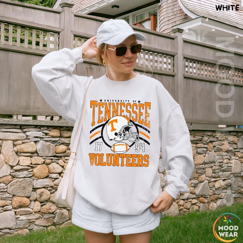 May include: A white long-sleeve sweatshirt featuring a graphic print that reads University of Tennessee Volunteers 1794 with an illustration of a football helmet and a football. The sweatshirt is paired with a white baseball cap and sunglasses.