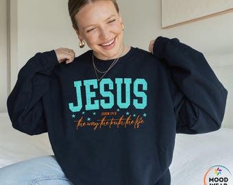Op de afbeelding: Zwart sweatshirt met het woord "JESUS" in turquoise blokletters. Onder het woord "JESUS" staat de tekst "JOHN 14:6" en "the way the truth the life" in oranje. Ronde hals.