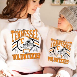 May include: Two white crewneck sweatshirts featuring a distressed orange and black graphic that reads University of Tennessee Volunteers, with a football helmet displaying a T logo and a football, plus the numbers 17 and 94 on the sides.