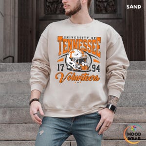 May include: Sand-colored sweatshirt featuring the text "UNIVERSITY OF TENNESSEE" in orange, along with a football helmet graphic. Below the helmet are the numbers "17" and "94", and the word "Volunteers".