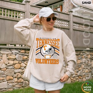 May include: A sand-colored crewneck sweatshirt featuring a graphic print that reads University of Tennessee 17 94 Volunteers, with an illustration of a football helmet and a football in the center, worn with a white baseball cap and sunglasses.