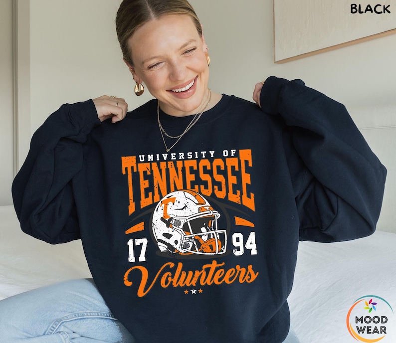 May include: A navy blue sweatshirt with orange and white graphics. The design features the words "University of Tennessee" and "Volunteers" along with a football helmet and the numbers "17" and "94". The sweatshirt is a crew neck style.