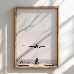 May include: A framed black and white print featuring a person sitting on a wooden pier by the water, with a vintage propeller airplane flying in the sky above. The artwork is enclosed in a light-toned wooden frame with a clean, rectangular border.