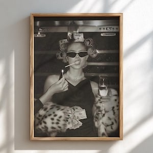 Può includere: Una stampa artistica incorniciata in bianco e nero che ritrae una persona con bigodini, occhiali da sole scuri e una stola di pelliccia. La persona tiene un fiammifero acceso, un calice di champagne e banconote da cento dollari in un ambiente scuro.