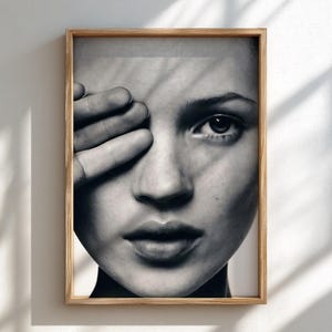 May include: A black and white framed portrait print featuring a close-up of a face with one hand partially covering the eye. The image displays high-contrast lighting with distinct shadows cast across the skin, housed in a light-toned wooden frame.