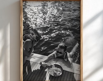 May include: A black and white framed photograph showing a person in sunglasses leaning over the edge of a boat deck, holding a fork and eating pasta from a bowl. Another person is partially visible nearby, reading a book by the water.
