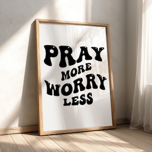Peut inclure: Une affiche murale rectangulaire dans un cadre en bois clair présentant le texte noir PRAY MORE WORRY LESS dans une police grasse, ondulée et rétro sur un fond blanc. Le design est centré dans le cadre, affichant une typographie nette.
