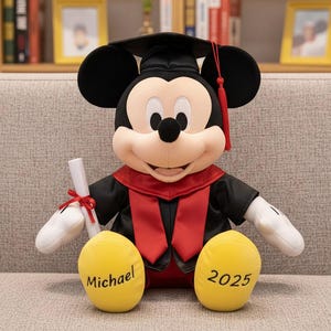 Peut inclure: Peluche de Mickey Mouse portant une tenue de remise de diplôme noire et rouge, coiffée d'un mortier. Il tient un diplôme enroulé, orné d'un ruban rouge. Les pieds jaunes portent les inscriptions "Michael" et "2025".