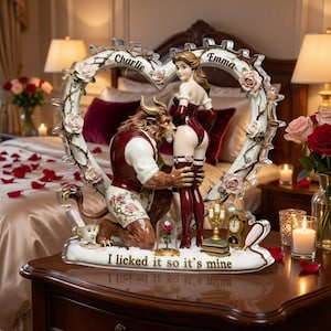 Op de afbeelding: Een romantisch beeldje met een beestachtig figuur en een vrouw in een hartvormige lijst versierd met rozen. De basis luidt "I licked it so it's mine". De namen "Charlie" en "Emma" staan op het frame. De figuren zijn in bordeauxrood en wit.
