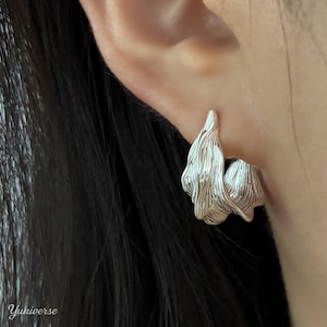 Può includere: Orecchini a cerchio color argento con un design strutturato e organico. Gli orecchini hanno un aspetto fluido e stratificato, simile a petali o metallo scolpito. Gli orecchini sono indossati su un orecchio.