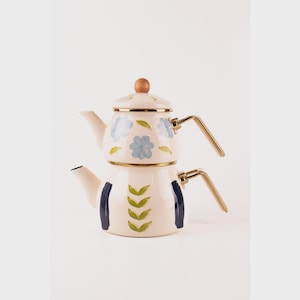 Puede incluir: Juego de tetera de esmalte color crema con diseño floral. La tetera superior presenta flores azules, mientras que la tetera inferior tiene hojas verdes y rayas azul marino. Tiene asas doradas y un pomo de madera en la tapa.