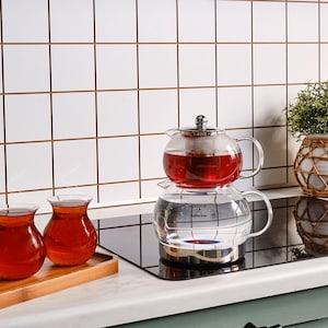 Puede incluir: Una tetera de vidrio transparente con tapa de acero inoxidable, llena de líquido rojo, apilada sobre una tetera de vidrio transparente más grande. Dos pequeñas tazas de té de vidrio llenas de líquido rojo se encuentran en una bandeja de madera.